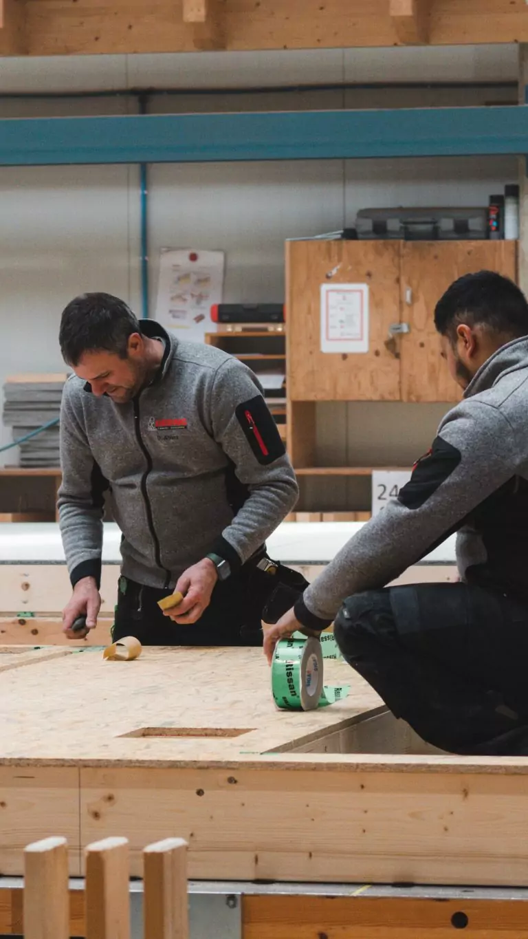 Zwei Männer in grauen Jacken arbeiten an einem Holztisch mit Werkzeug und Klebeband in einer Werkstatt.