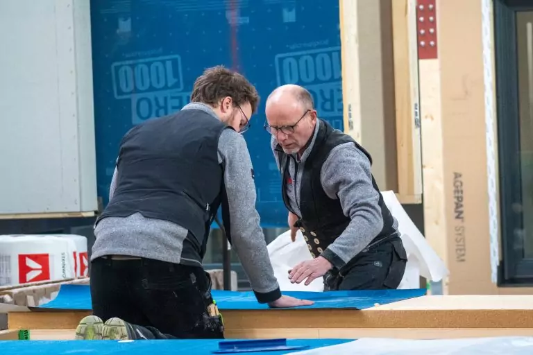 Zwei Handwerker arbeiten konzentriert an einem Tisch mit blauer Folie im Freien, umgeben von Baumaterialien.
