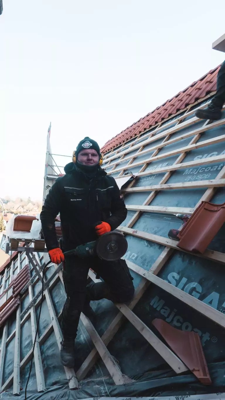 Arbeiter mit Ohrenschützern und Flex arbeitet auf einem Ziegeldach, umgeben von Baugerüst und Dachziegeln.