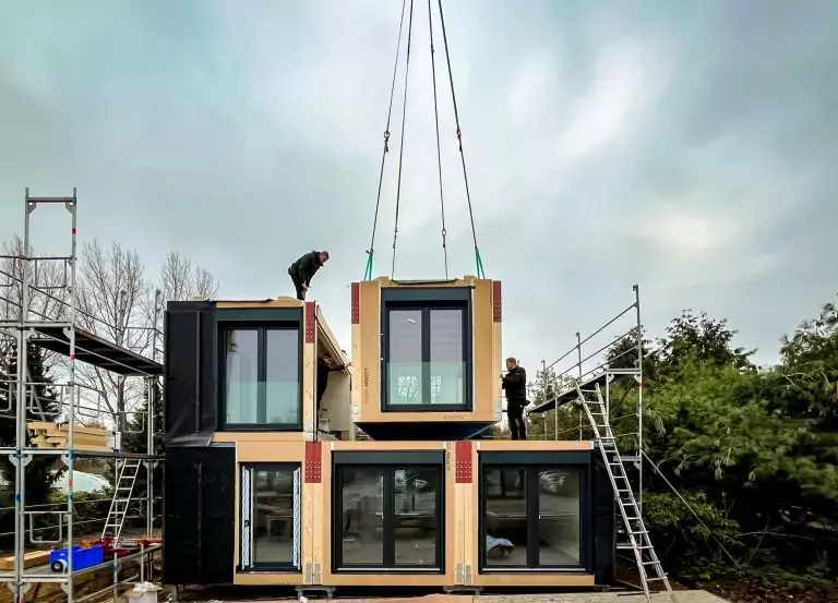 Modulares Haus wird montiert; Arbeiter auf einem Gerüst, während ein weiteres Modul mit einem Kran platziert wird.