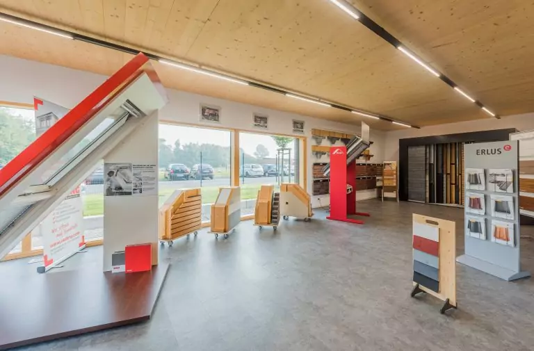 Ausstellungsraum mit Dachmaterialmustern, Holzvertäfelung und großen Fenstern, die auf einen Parkplatz blicken.
