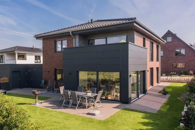Modernes zweistöckiges Haus mit roter Ziegelfassade, Terrasse und Garten bei sonnigem Wetter.
