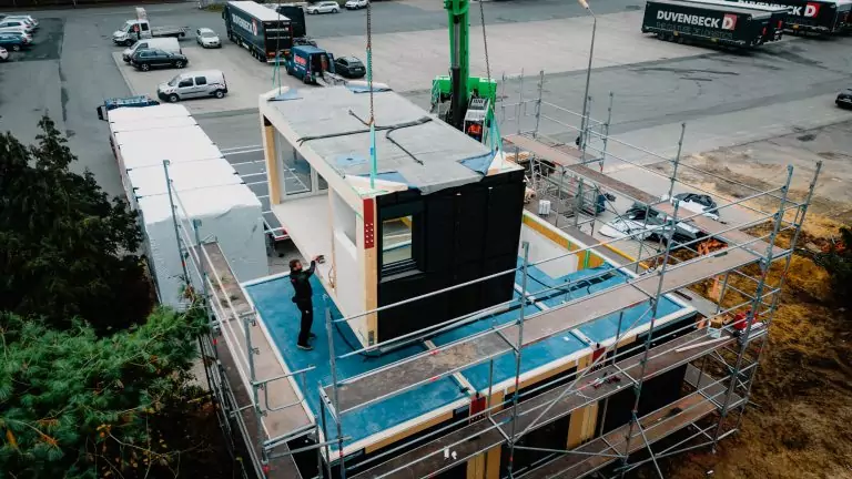 Bauarbeiter montiert ein Gebäudeteil mit einem Kran auf einer Baustelle, umgeben von Gerüst und parkenden Autos.