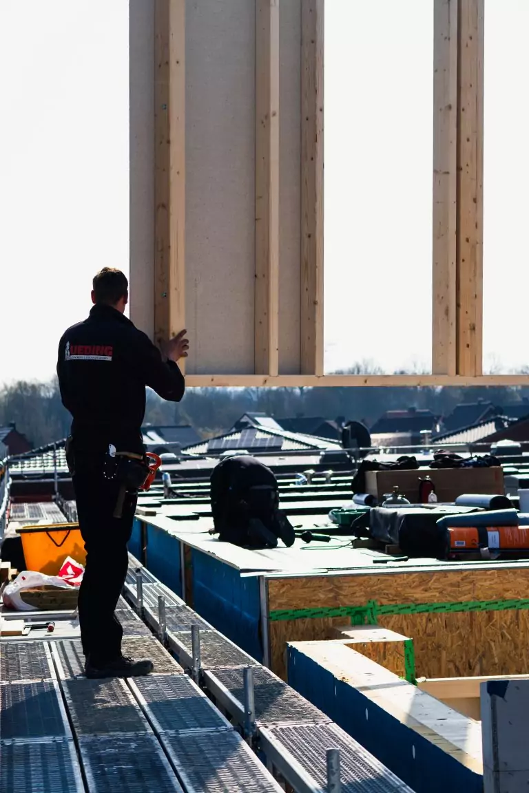 Ein Bauarbeiter montiert eine Holzplatte auf einem Dachgerüst, ein weiterer Arbeiter kniet in der Nähe des Materials.