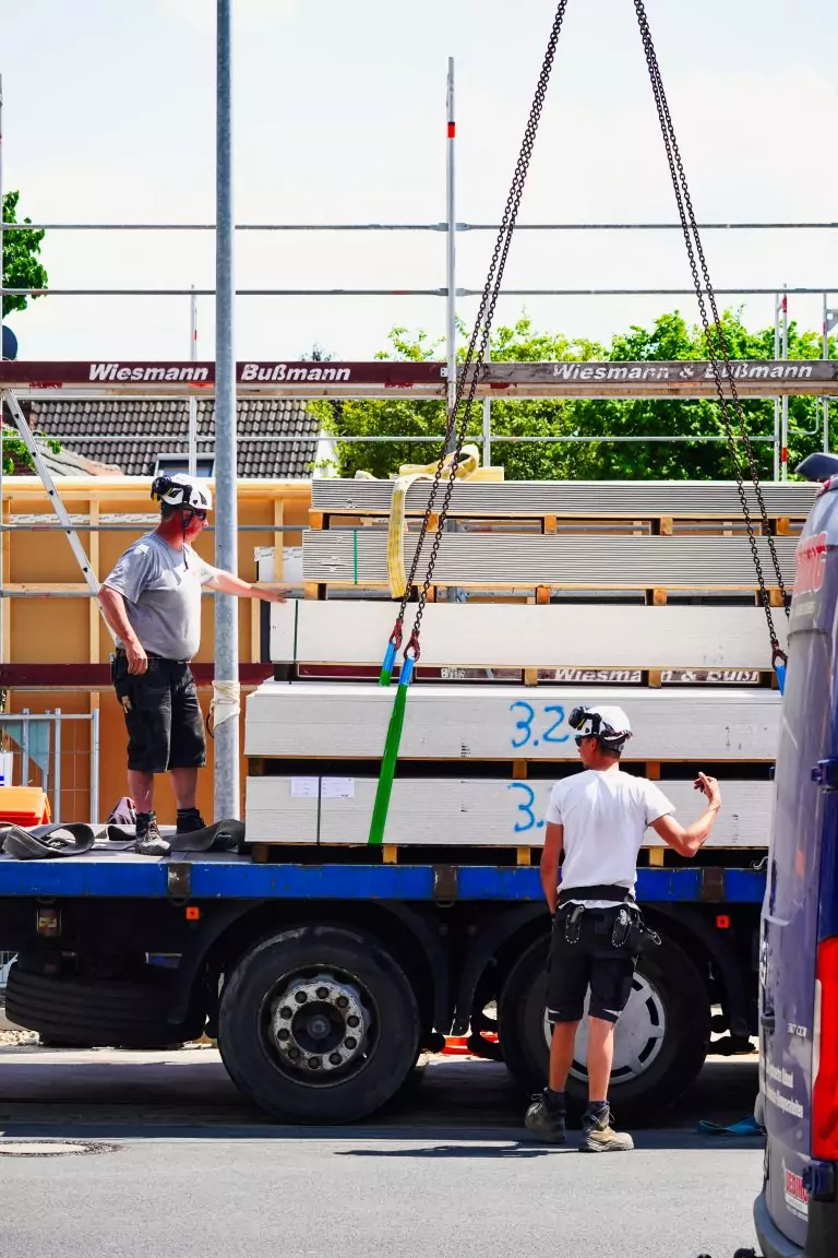 Zwei Bauarbeiter laden Holzplatten von einem LKW ab, unterstützt von einem Kran vor einem Gerüst.