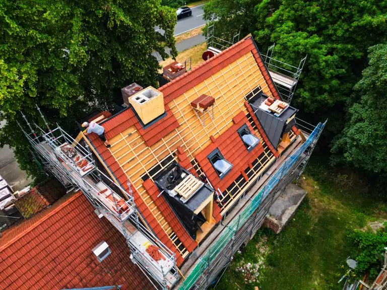 Dacharbeiten: Ein Bauarbeiter verlegt Ziegel auf einem eingerüsteten Dach neben Bäumen und einer Straße.