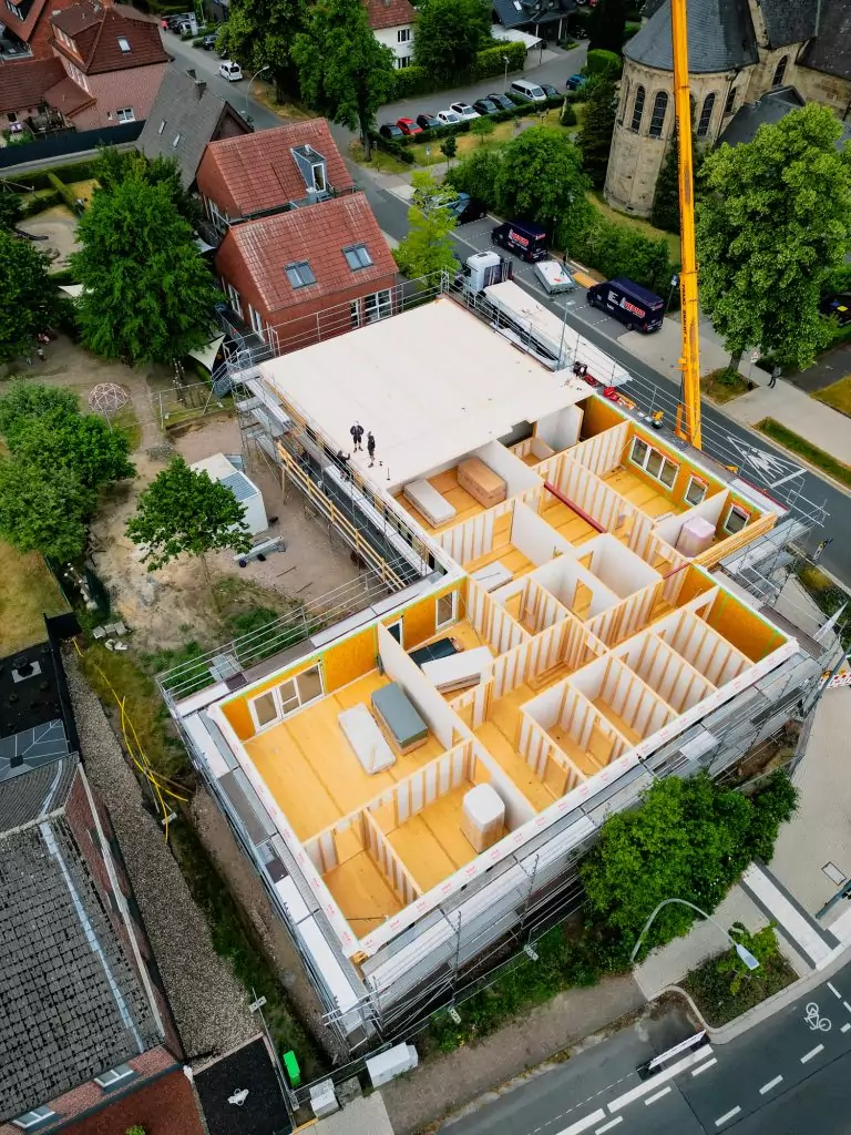 Vogelperspektive eines Gebäudes im Rohbau mit sichtbarem Grundriss, umgeben von Bäumen und einer Straße.