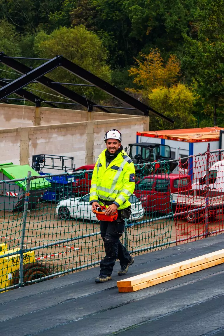 Bauarbeiter in gelber Jacke und Helm steht auf Baustelle mit Maschinen und Zäunen im Hintergrund.