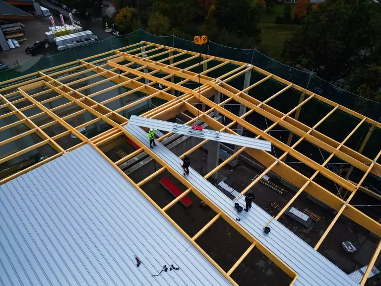 Arbeiter montieren Metallpaneele auf einem großen Holzrahmendach einer Baustelle aus der Vogelperspektive.