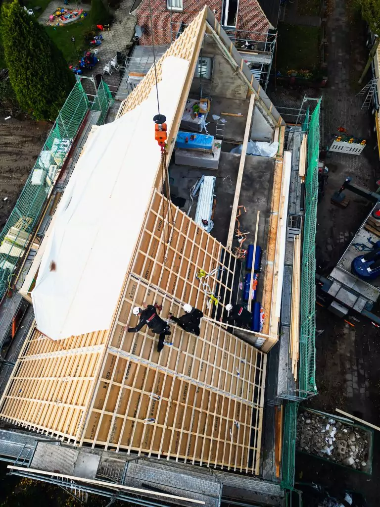 Bauarbeiter errichten ein Dach mit Holzkonstruktion, gesichert mit Kran, in einem Gebäudebauprojekt aus der Vogelperspektive.