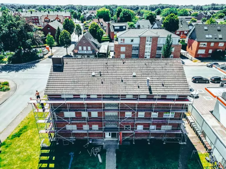 Rotes Backsteinhaus mit Gerüst, Straße und geparkten Autos im Vordergrund, Wohngebiet mit Bäumen im Hintergrund.