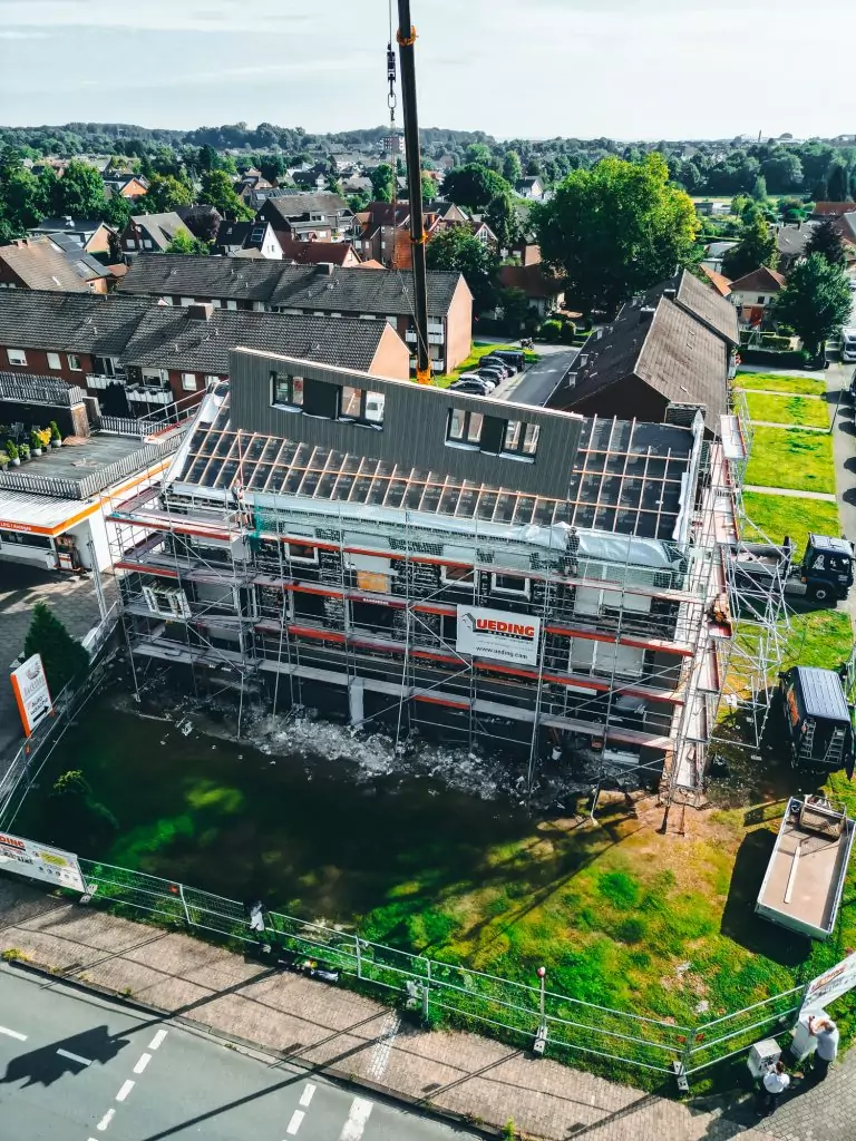 Kran hebt Gebäudeteil auf Baustelle in städtischer Umgebung. Gerüst um Gebäude, grüne Vegetation im Hintergrund.