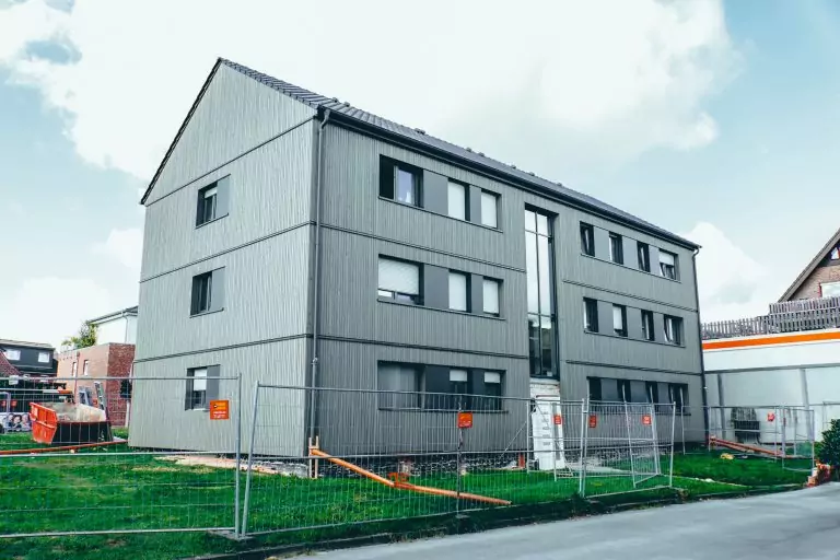 Graues dreistöckiges Gebäude mit Bauzaun davor. Im Hintergrund blauer Himmel und andere Gebäude.