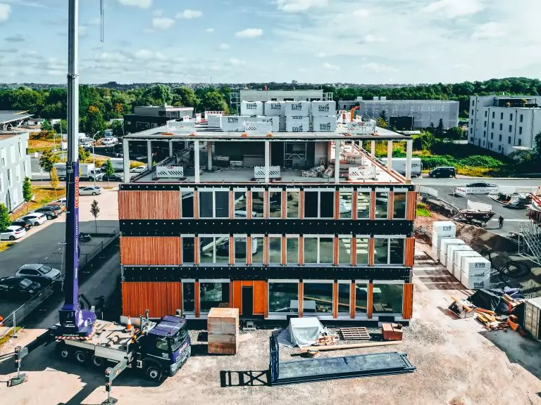 Gebäude im Bau mit Kran, Teilverglasung, umgeben von Grünflächen und parkenden Autos bei sonnigem Wetter.