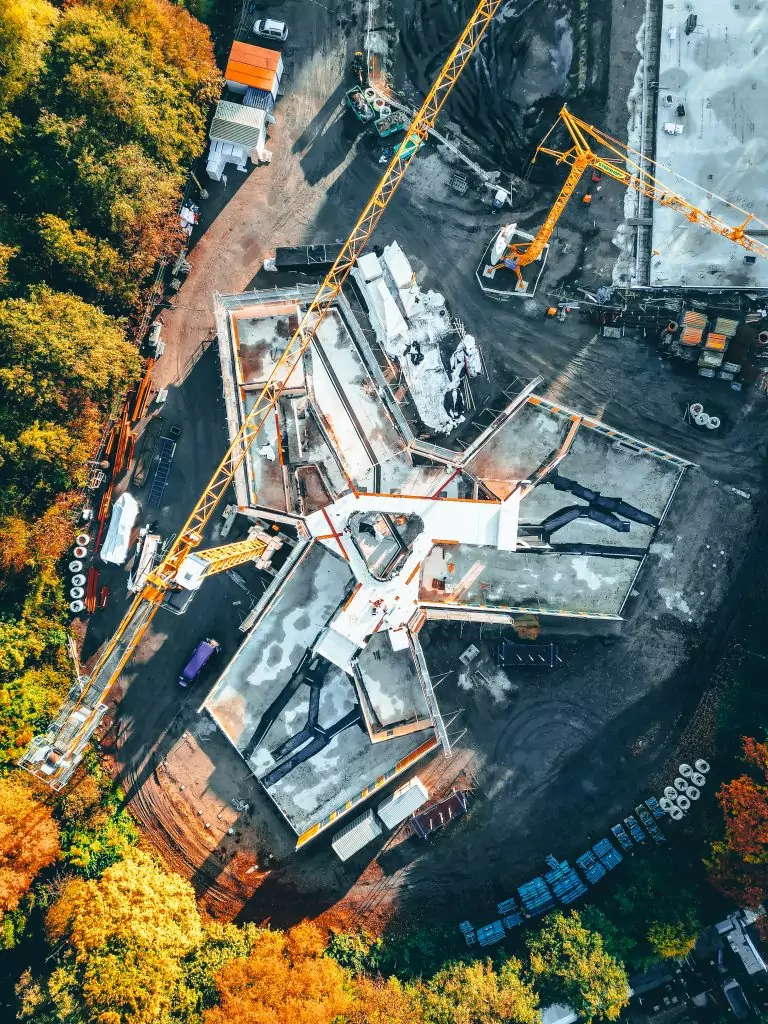 Luftaufnahme einer Baustelle im Wald, mit Kränen und Gebäudeteilen in herbstlicher Umgebung.