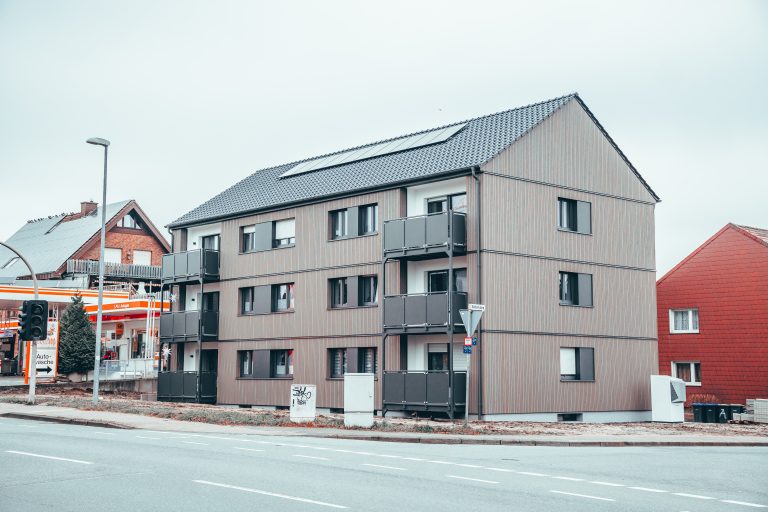 Mehrfamilienhaus mit Solarpanelen auf dem Dach neben einer Tankstelle an einer Straße.